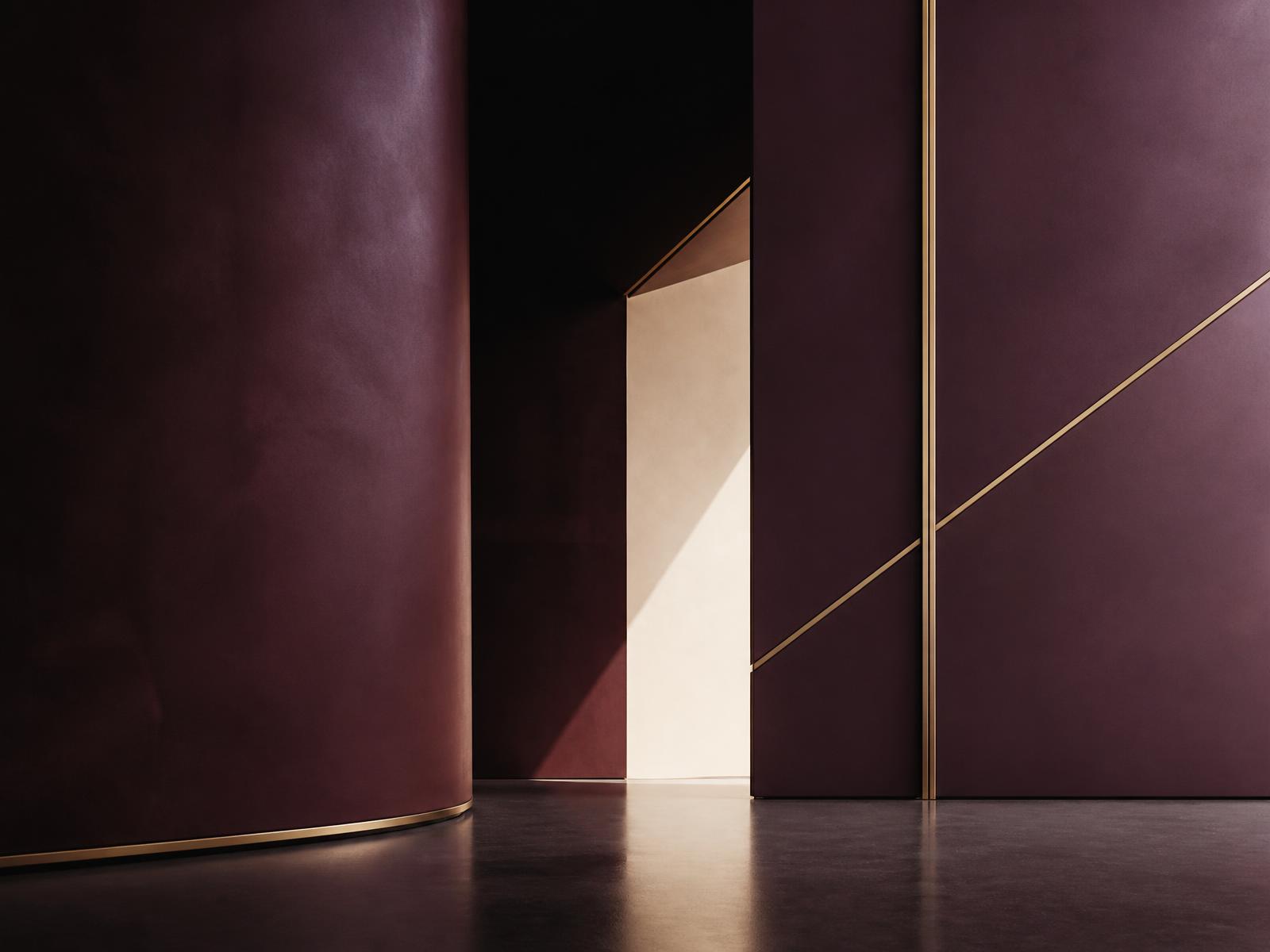 A deep plum architectural interior where a curved wall and thin gold lines frame a sunlit opening.
